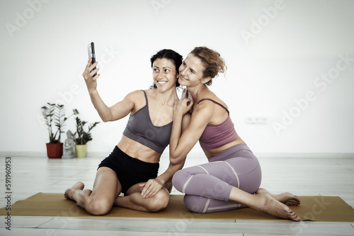 Two fitness women friend sitting on mat together and taking selfie on phone indoors