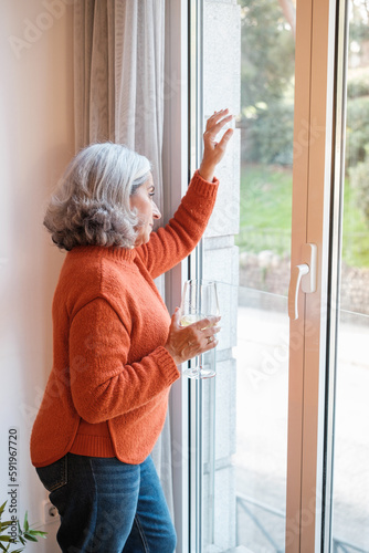 Senior woman with white hair having a glass of wine while relaxing looking out the window at home. Concept: Lifestyle, relax, disconnection