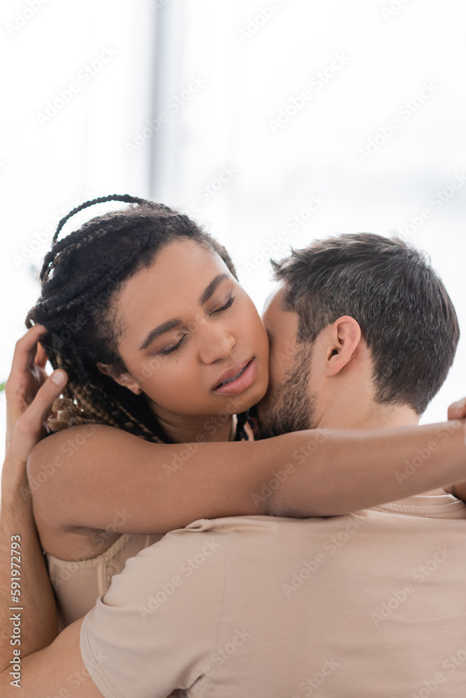 sexy african american woman with closed eyes embracing brunette man at home.