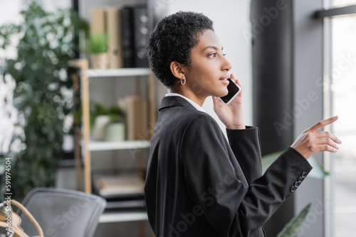 side view of young african american american businesswoman in black blazer pointing with finger and talking on cellphone in office.