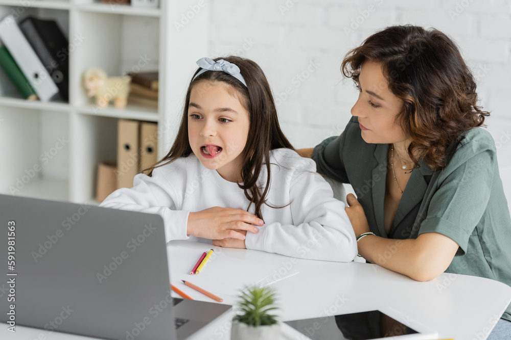 Parent sitting near daughter having speech therapy online lesson near devices at home. Parent sitting near daughter having speech therapy online lesson near devices at home.