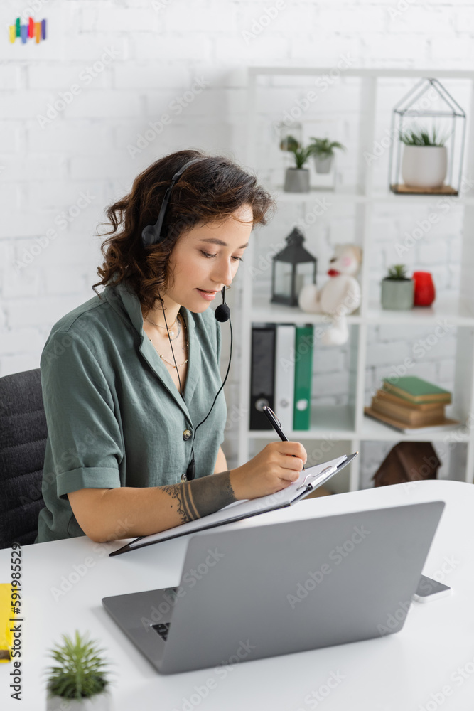Speech therapist in headset writing on clipboard near laptop in consulting room. Speech therapist in headset writing on clipboard near laptop in consulting room.