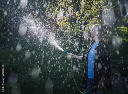 Beautiful young woman having fun in summer garden with garden hose splashing rain. Image with soft focus and bokeh