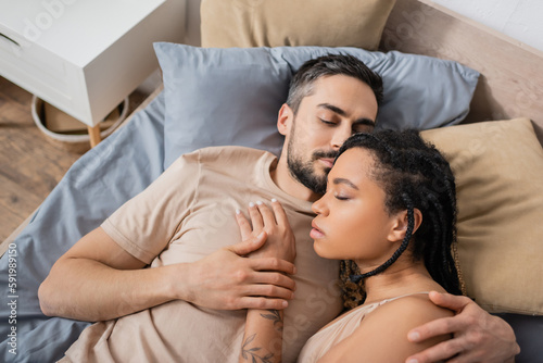 top view of bearded man with closed eyes hugging sensual african american woman while lying on bed at home.