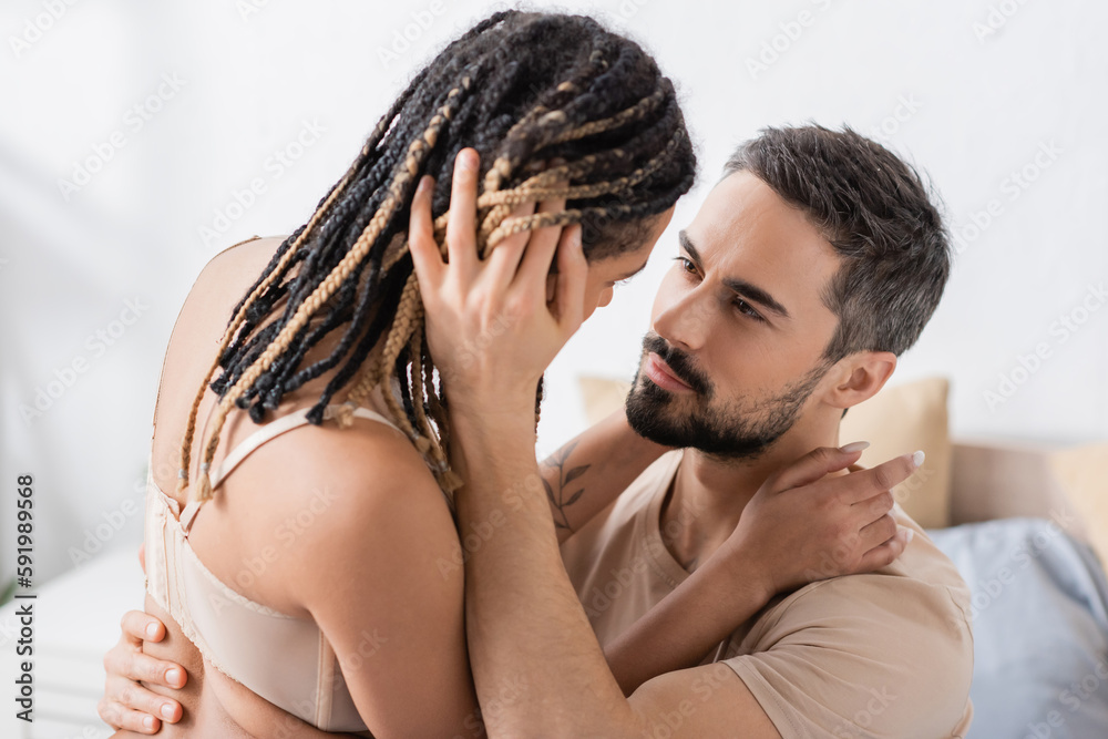 Fototapeta premium brunette bearded man embracing sexy african american woman with dreadlocks in bedroom at home.