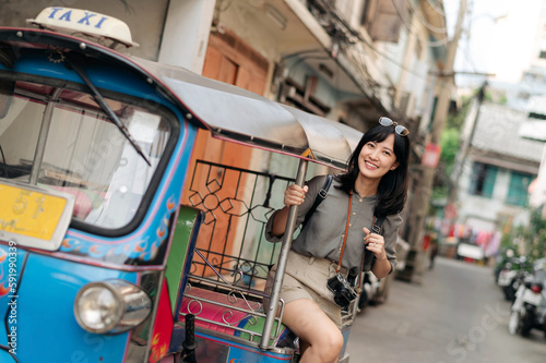 Young Asian woman backpack traveler standing a side of Tuk Tuk taxi on summer vacations at Bangkok, Thailand.