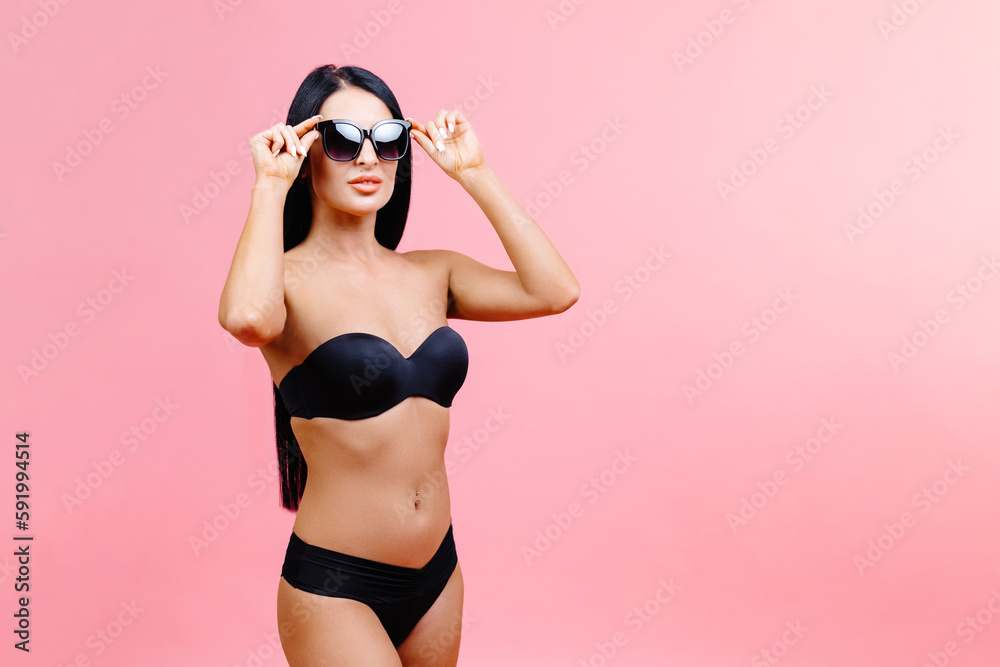 Young woman in summer black swimsuit and sunglasses standing on pink background in studio Young woman in summer black swimsuit and sunglasses standing on pink background in studio