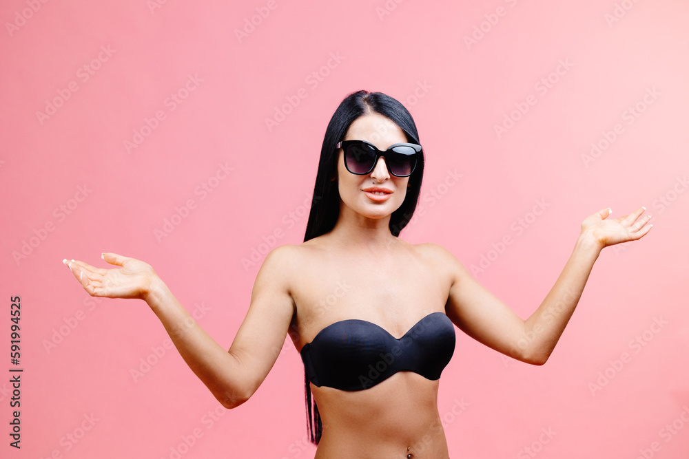 Young woman in summer black swimsuit and sunglasses standing on pink background in studio