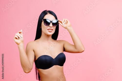 Young woman in summer black swimsuit and sunglasses standing on pink background in studio