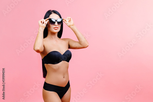 Young woman in summer black swimsuit and sunglasses standing on pink background in studio