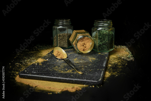 Three kinds of herbs or spices in glass jars sitting in a row on a black background with a spoon and dustings of herbs and spices on the countertop; Studio photo