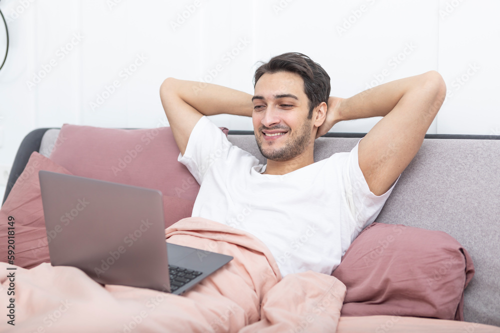 Handsome smiling man lying on the bed and working on laptop in the bedroom Handsome smiling man lying on the bed and working on laptop in the bedroom