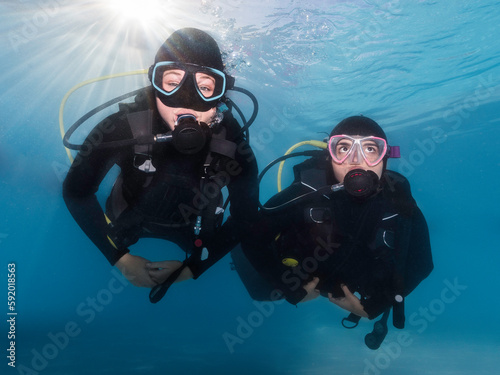 Closeup of two scuba divers underwater swimming towards the camera with the bright sunrays shining through the water surface behind them