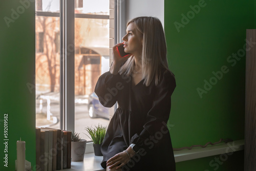 a young blonde woman in a black dress sits on the windowsill in the office, talking on the phone and looking out the window