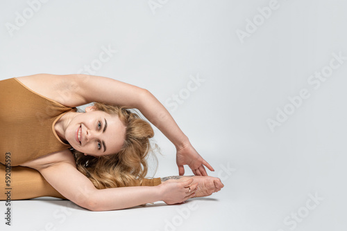Young beautiful curly-haired barefoot woman lying on the floor and stretching her body. Long-haired smiling athlete warming up and looking at the camera