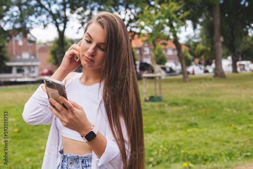 Attractive brunette long hair woman wear white top and shirt hold mobile phone and chatting, messaging, look serious and calm. Amazing woman walk in the park, touch her earphones girl answer to call.