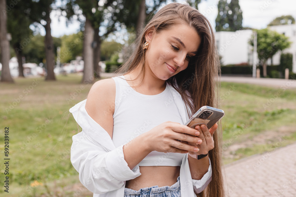Attractive brunette long hair woman wear white top and shirt hold mobile phone and chatting, messaging, look happy and smiling. Amazing enjoy woman walk in the park, girl look at phone. Attractive brunette long hair woman wear white top and shirt hold mobile phone and chatting, messaging, look happy and smiling. Amazing enjoy woman walk in the park, girl look at phone.