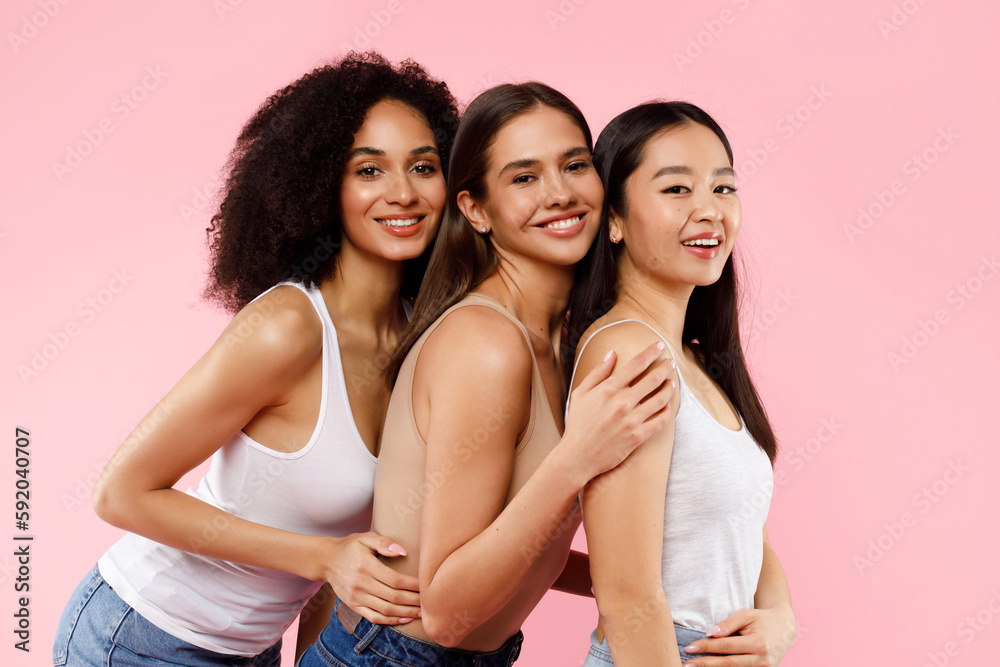 It girls. Happy three multiracial young ladies in casual embracing, posing and smiling at camera on pink background