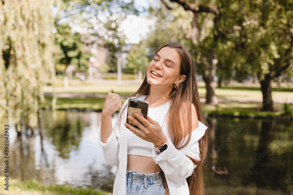 Delighted brunette girl making winner gesture and using mobile phone walking on the park near river. Portrait of surprised brunette girl surfing internet on mobile phone outdoors, woman in shirt, top.