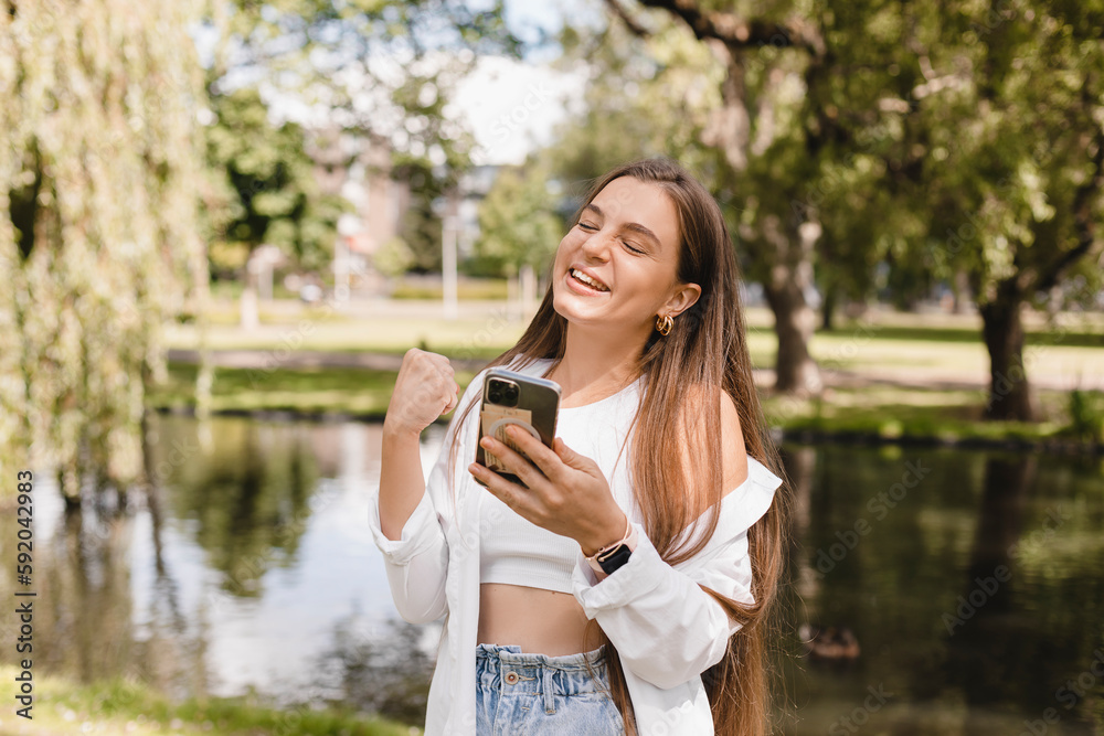 Delighted brunette girl making winner gesture and using mobile phone walking on the park near river. Portrait of surprised brunette girl surfing internet on mobile phone outdoors, woman in shirt, top. Delighted brunette girl making winner gesture and using mobile phone walking on the park near river. Portrait of surprised brunette girl surfing internet on mobile phone outdoors, woman in shirt, top.