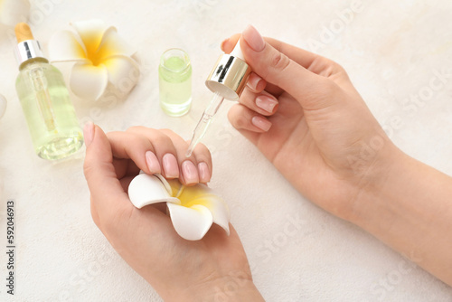 Female hands with bottle of cuticle oil and plumeria flowers on light background, closeup