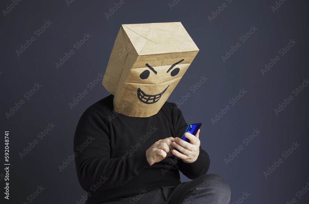 A man with a bag with an evil smiley on his head is talking on the phone. A man with a bag with an evil smiley on his head is talking on the phone.
