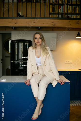Gorgeous business lady in beige pantsuit sitting on table in kitchen. Stylish kitchen interior design, working lifestyle photo