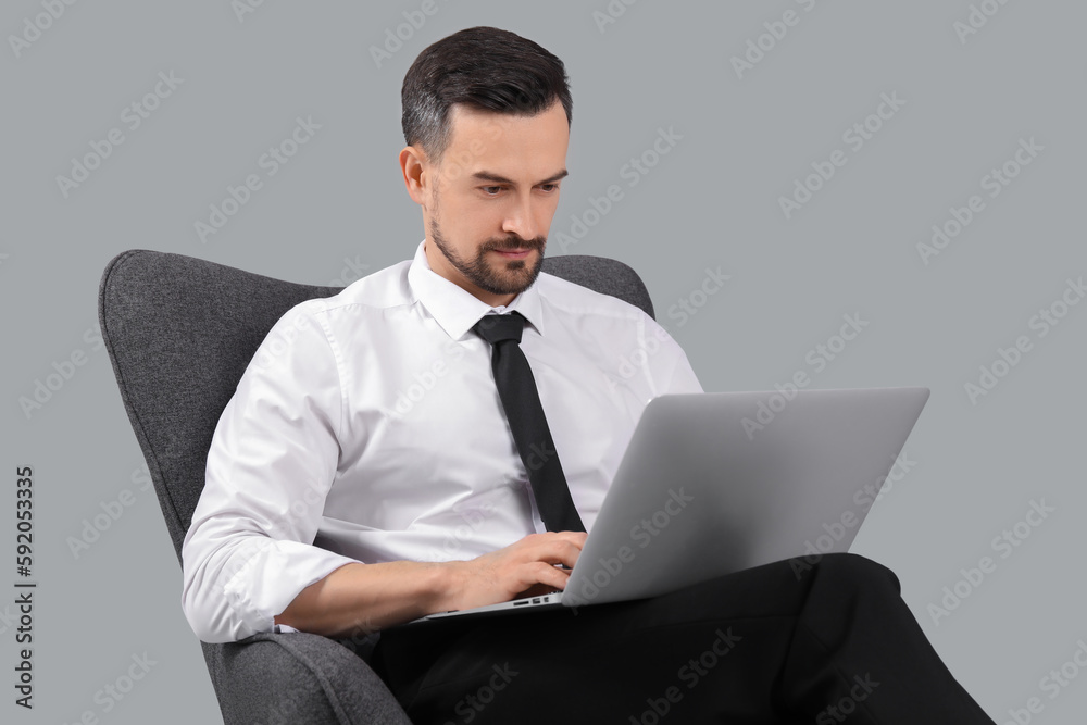 Handsome businessman working with laptop in armchair on grey background