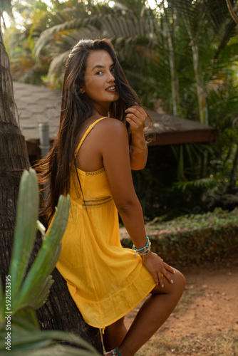 Smiling flirting brunette woman in yellow dress lean on palm tree, look camera and touch hair in green jungle plants