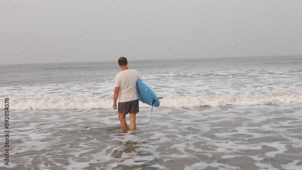 Rearview calm mature man going to sea waves and turning to camera, carrying surfboard against gray sky. Beach travelling