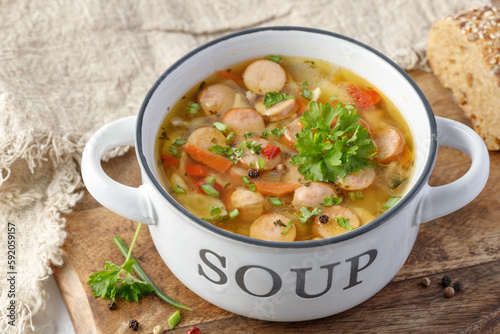 Fresh hot soup with meat sausage slices, beans, carrot, pasta and green herbs in bowl on wooden board.