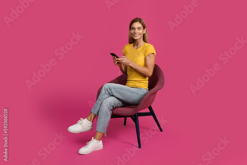 Glad cheerful middle aged european blonde lady sitting in chair, typing on smartphone © Prostock-studio