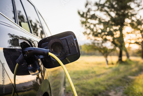 Black electric car with the power cable supply plugged in, in the blurred background visible green area and trees. Eco friendly and renewable energy concept.