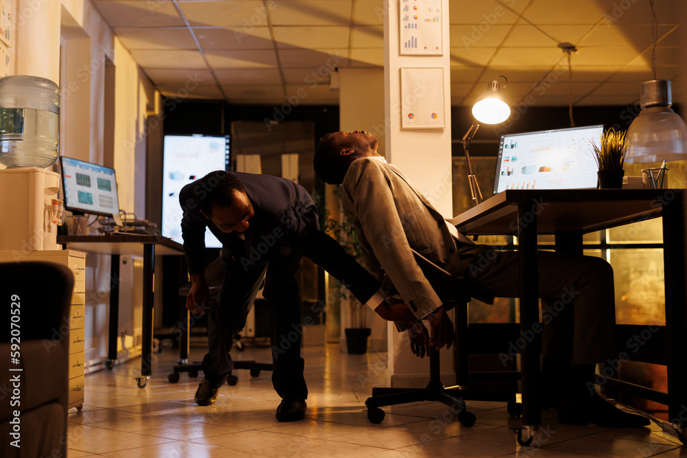 Manager pulls colleagues chair after falling asleep on chair in startup office, african american employees working overhours at marketing strategy analyzing company statistics. Business concept