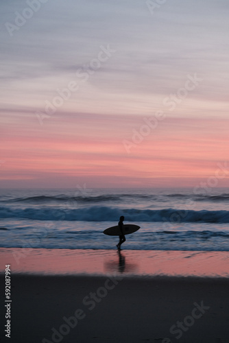 vertical photography of a silhouette of a surfer with a surfboard walking on the beach during sunset