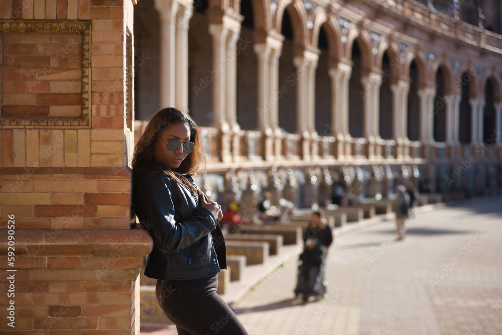 Obraz premium Young, beautiful, Latin and South American woman with leather top and jacket, sunglasses and jeans, leaning against a brick wall, posing sensual and attractive. Concept beauty, fashion, diversity.