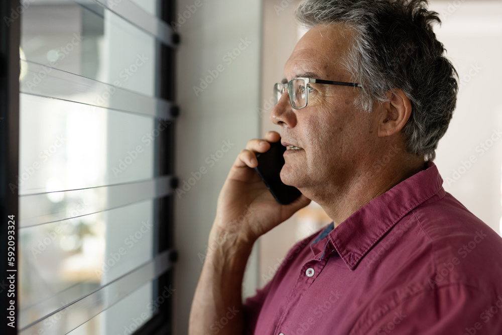 Caucasian senior man talking over smartphone and looking through window while standing at home Caucasian senior man talking over smartphone and looking through window while standing at home