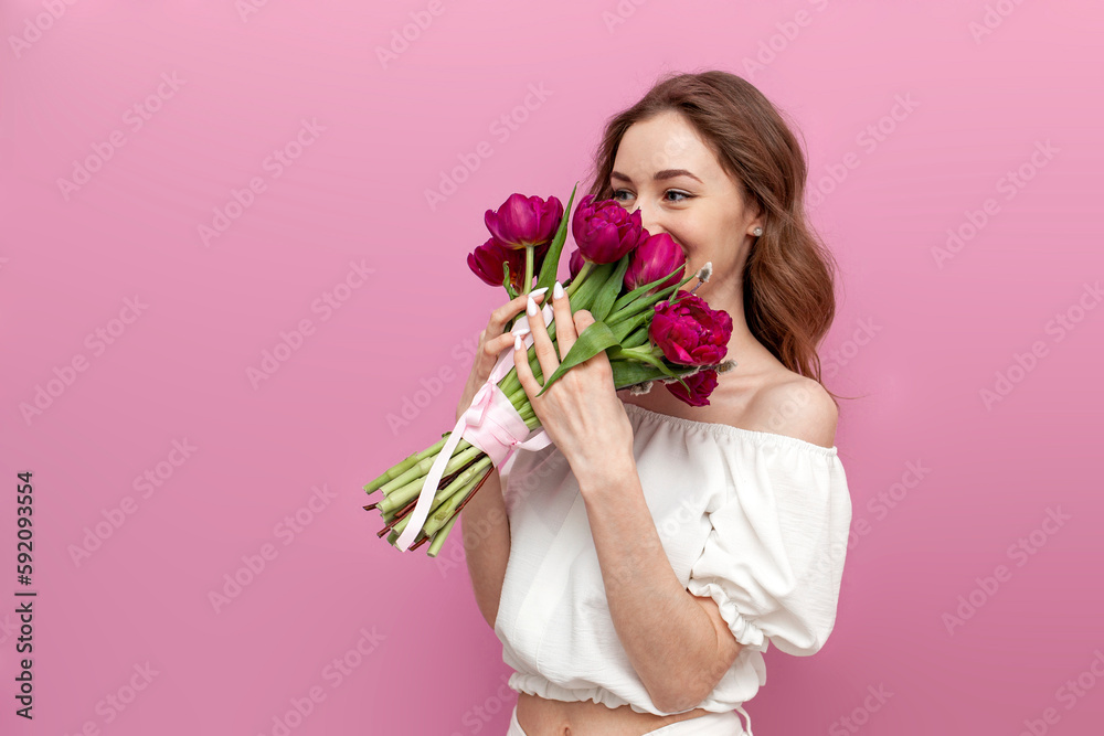 cute young woman in festive outfit holds bouquet of pink tulips and smells them on pink isolated background