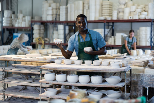 Positive african-american man potter in apron standing in ceramic workshop, holding crafted cups of bowls, looking at camera and smiling.