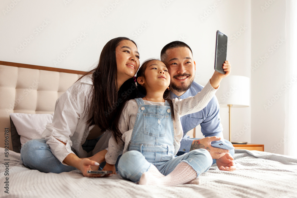 asian family sit on the bed and communicate via video call, little korean girl with mom and dad takes selfie
