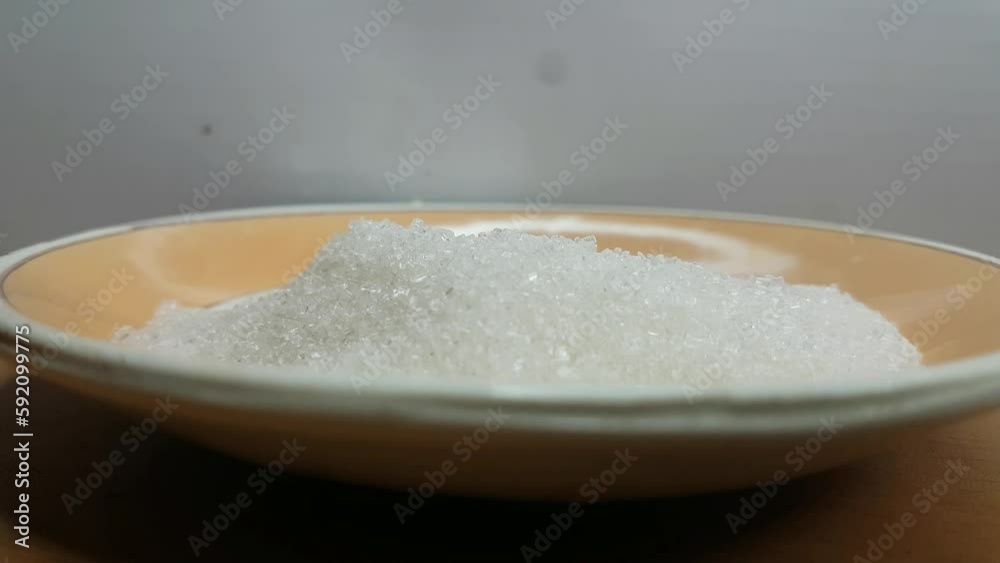 Pile of refined and powdered white sugar crystals inside a ceramic tea bowl or plate with plastic spoon placed on top of it. High sugar consumption in India and Asia concept. Close up macro side view. Pile of refined and powdered white sugar crystals inside a ceramic tea bowl or plate with plastic spoon placed on top of it. High sugar consumption in India and Asia concept. Close up macro side view.