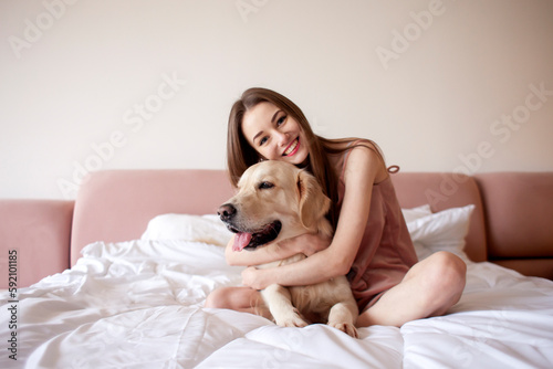 cute girl in pajamas sits on the bed and hugs golden retriever breed dog, woman in the morning at home with pet