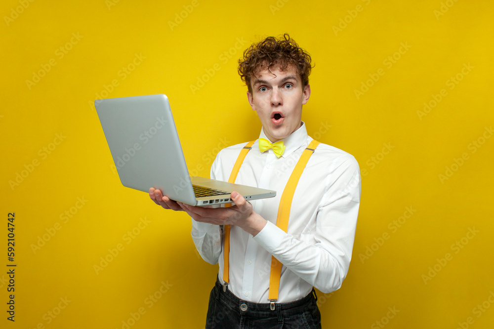 shocked guy in festive outfit uses laptop and is surprised on yellow isolated background, student in bow-tie shirt