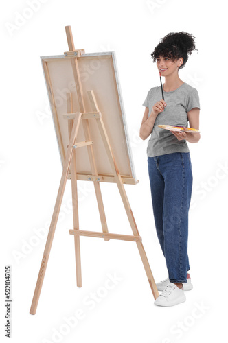 Young woman holding brush near easel with canvas against white background