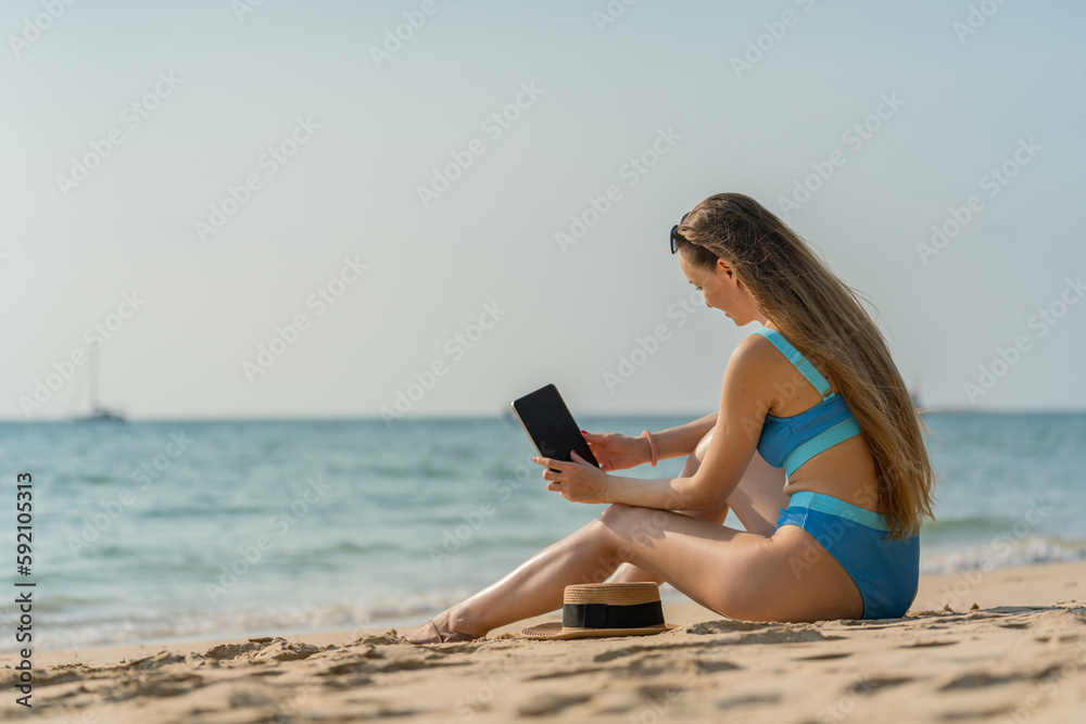 Beautiful caucasian woman using digital tablet at the beach. Female digital nomad working from the beach using her digital tablet. Sexy white woman in bikini. Workcation.