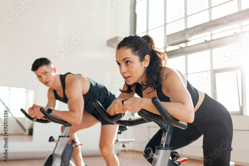 athletic couple of cyclists train on static bicycle simulator, man trainer and fitness woman go in for sports together