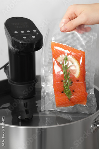 Woman putting vacuum packed salmon into pot with sous vide cooker in kitchen, closeup. Thermal immersion circulator photo