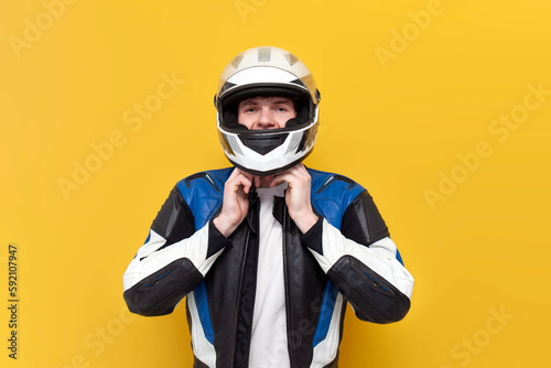 young guy motorcyclist in leather jacket puts on helmet on yellow isolated background