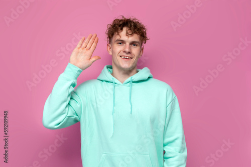 curly guy in mint hoodie greets and waves his hand on pink isolated background, man says goodbye and shows his palm