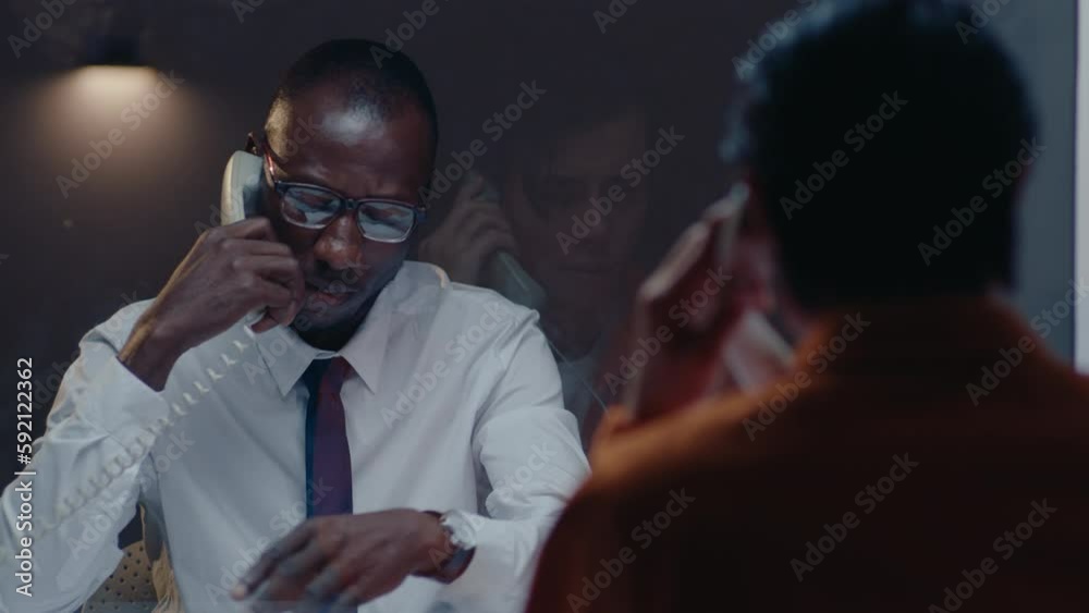 Black detective showing mugshots of suspects through the glass wall, questioning prisoner on the phone in the visiting room, investigating crime